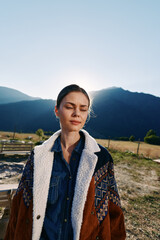 Fototapeta premium Woman portrait in mountains with sunlight behind her, eyes closed and serene expression, wearing warm patterned coat and denim shirt in outdoor nature with soft backlight.