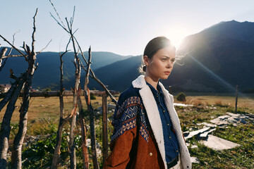 Naklejka premium Woman portrait in countryside mountains with warm sunlight wearing a wool coat by a rustic fence, thoughtful young traveler captured in golden backlight and scenic valley landscape.