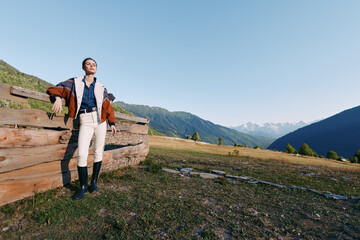 Fototapeta premium Man leaning on wooden fence in mountains meadow wearing stylish coat and boots, countryside portrait with wide sky and scenic landscape, fashion pose and relaxed outdoors vibe.