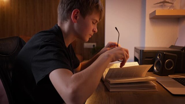 Tired teenager experiencing eye strain and exhaustion while study book at desk in late hours of night. Boy removes glasses, massages weary eyes, and lets out yawn