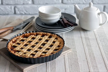 Freshly baked lattice pie on a rustic table beside stacked plates, bowls, and a white teapot, creating a cozy homemade dessert scene.