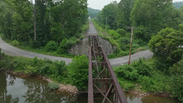 An abandoned railway bridge stretches over a calm river. Dense trees and vegetation cover the area, creating a serene and peaceful atmosphere.