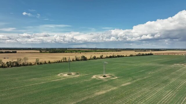 Fast half-circle drone flight over fields under stormy clouds, with a high-voltage line on one field and wind turbines on another, highlighting dynamic landscape and energy infrastructure.