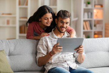 Happy Indian couple using digital tablet while resting on couch at home, shopping online, browsing internet or ordering food in app