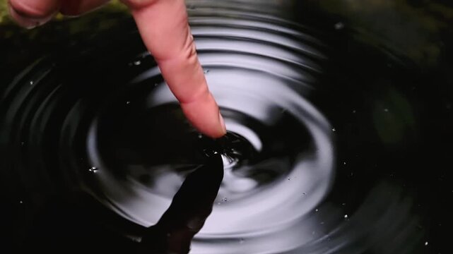 Close-up of finger touching dark water surface creating ripples