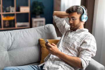 Obraz premium Relaxed Indian man chilling on couch at home, using wireless headphones and cellphone, watching video content online, copy space