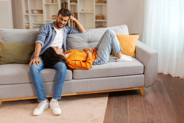 Happy Indian spouses spending free time together, resting on couch at home in living room, woman...
