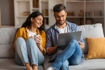 Happy Indian spouses sitting on sofa and browsing laptop together, shopping online, choosing movie or surfing Internet, woman holding cup with coffee