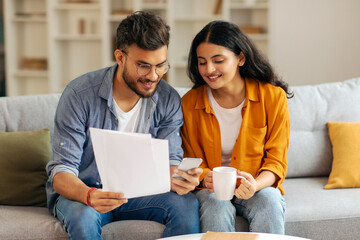 Indian couple intently examining documents together, with cellphone, woman holding coffee mug, sitting on couch in living room