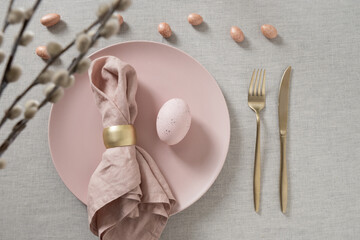 Minimal Easter table setting with pink plates, pastel eggs,willow branches, linen napkin and golden cutlery. Elegant spring holiday table decor with copy space and neutral background.