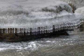 Icicles hang delicately from a snow-covered rock as a stream flows rapidly below. The contrast between the icy formations and the moving water creates a dynamic scene.
