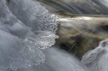 Icicles hang delicately from a snow-covered rock as a stream flows rapidly below. The contrast between the icy formations and the moving water creates a dynamic scene.