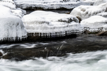 Icicles hang delicately from a snow-covered rock as a stream flows rapidly below. The contrast between the icy formations and the moving water creates a dynamic scene.