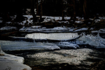 A serene winter scene captures a stream partially frozen, surrounded by snow-covered forest. Icicles hang from the edges of the ice.