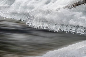 Icicles hang over a flowing river, capturing the contrast between the stillness of the ice and the movement of the water. The scene evokes a sense of tranquility and cold beauty.
