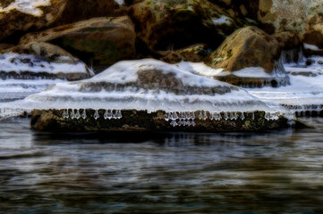 Icicles hang over a flowing river, capturing the contrast between the stillness of the ice and the movement of the water. The scene evokes a sense of tranquility and cold beauty.