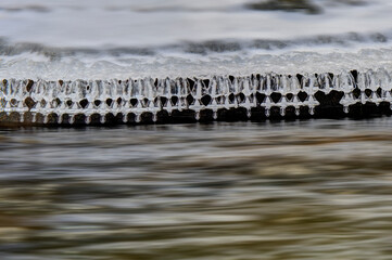 Icicles hang over a flowing river, capturing the contrast between the stillness of the ice and the movement of the water. The scene evokes a sense of tranquility and cold beauty.