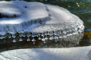 A natural winter scene featuring a row of teardrop-shaped icicles hanging from a snow-covered surface above a body of water. Some of the frozen forms display dark inclusions or reflections 
