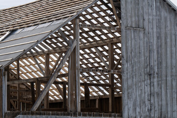A partially constructed or deteriorated wooden barn stands in a rural setting, its lattice of beams exposed where walls and roof sections are missing. One portion of the roof is covered 