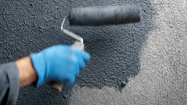 Close-up of a gloved hand rolling dark paint onto a textured gray wall, catching light in warm glow!!!