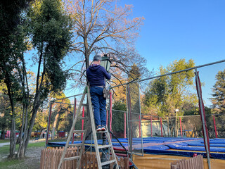 Caucasian man standing on a ladder repairing a lamppost in an urban public park. The scene illustrates maintenance work, use of equipment, and organized setup in a public outdoor space. © quietmind_art