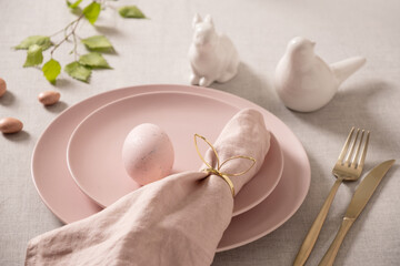 Elegant Easter table setting with pastel pink ceramic plate, gold cutlery, linen napkin and decorative egg. Minimal spring holiday dining concept with soft natural light.