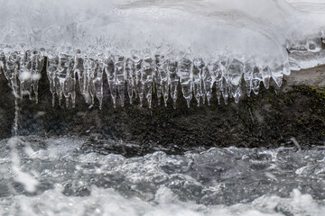 Icicles hang delicately from a snow-covered rock as a stream flows rapidly below. The contrast between the icy formations and the moving water creates a dynamic scene.