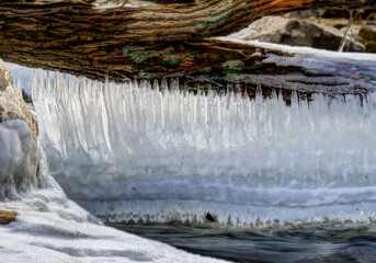 A serene winter scene featuring a fallen tree trunk suspended over a partially frozen stream. Long, delicate icicles hang from the underside of the trunk, forming a shimmering curtain 