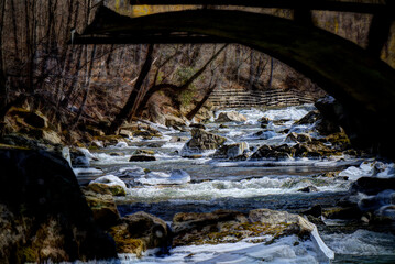 A scenic winter view of a rocky stream rushing through a forested landscape. Whitewater rapids cascade over ice-covered rocks, while leafless trees line the banks in quiet stillness. 