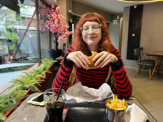Teenage Caucasian female with red hair and striped sweater wearing glasses eats a burger at a cafe showing casual dining, table setup, and public seating arrangement in an everyday environment.