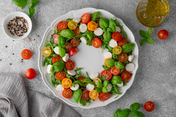 Caprese salad with arugula, mozzarella, cherry tomatoes, fresh basil and balsamic glaze on a white plate on a gray concrete background. Top view, copy space.