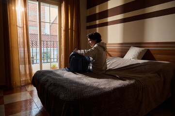 Fototapeta premium Young Man Packing Backpack on Bed in a Sunlit Hotel Room by a Balcony Window