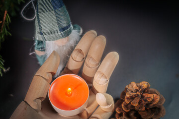 A wooden mannequin's hand holds an orange tealight surrounded by pine cones, green foliage, and a touch of festive fabric decor.  A warm, festive composition.