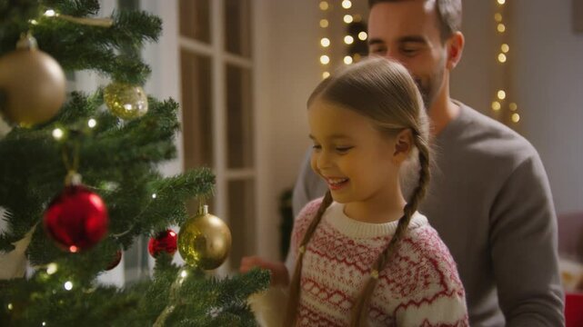 Familia joven decorando un &aacute;rbol de Navidad en casa, padres y ni&ntilde;a peque&ntilde;a compartiendo un momento feliz y cercano. Escena c&aacute;lida y acogedora con luces navide&ntilde;as, adornos rojos y ambiente hogare&ntilde;o