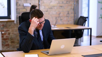 video. Caucasian man is working in office. Woman is sitting in background. Businessman is sitting on the table, reading documents, taking off his glasses, looks very tired.