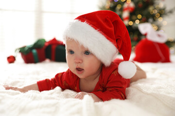 Cute baby in Santa hat on bed against blurred lights indoors. Christmas season
