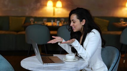 Smiling businesswoman sitting in restaurant while talking online while using laptop, waving hand. Woman looking on screen, waving hand. Business, lifestyle concept.