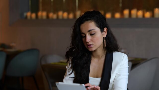 Young businesswoman holding digital tablet while tapping on the screen, drinking coffee while sitting in restaurant. Business, lifestyle concept. - Powered by Adobe
