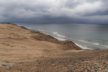 sand dunes, eroded coastal cliffs, calm, grey sea, heavy, overcast skies. moody shoreline landscape, sense of solitude raw natural beauty, wilderness, erosion, dramatic coastal environments.