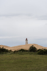 a solitary lighthouse atop golden sand dunes, set against overcast sky, calm, atmospheric coastal scene. costal vegetration green bushes in forgreground