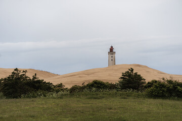 a solitary lighthouse atop golden sand dunes, set against overcast sky, calm, atmospheric coastal scene. costal vegetration green bushes in forgreground