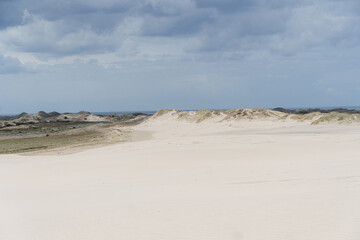 A wide coastal dune landscape with smooth white sand, rolling hills, and distant grasslands beneath a moody, cloud-filled sky, evoking tranquility and natural openness.