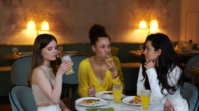 Young girlfriends having dinner in a restaurant while eating bruschettas and drinking a cocktails, gossiping together. Meeting of friends. Lifestyle, friendship concept. Slow motion