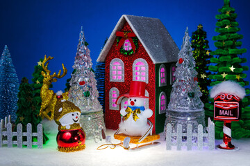 A snowman and a snowman in a red hat on a sled among glass Christmas trees near a red Christmas house surrounded by a fence