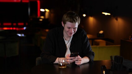 Happy young man sitting in a restaurant and drinking whiskey while typing a message on the mobile phone. Lifestyle concept.