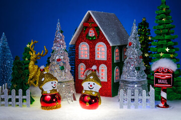 Two snowmen among glass Christmas trees near a red Christmas house surrounded by a fence