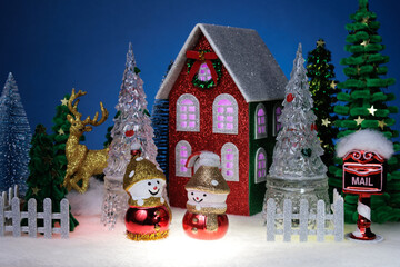 Two identical snowmen among glass Christmas trees near a red Christmas house surrounded by a fence
