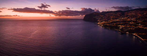 Atlantic waves meet rugged Madeira cliffs as Funchal lights glow down hills. Orange and magenta...