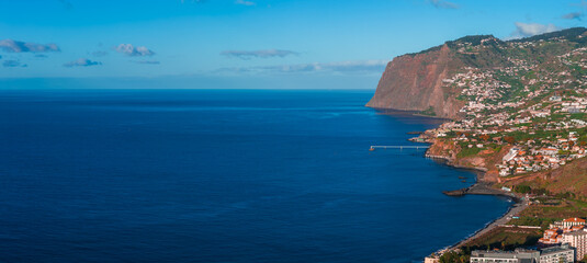 Wide coastal panorama of Madeira near Funchal shows Cabo Girao, terraced towns, whitewashed houses, a small pier, and a winding coastal road in soft afternoon light. © Aerial Film Studio