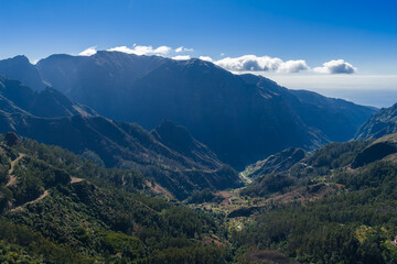 Fototapeta premium Aerial view shows Madeira central mountains near Pico do Arieiro, terraced slopes, laurel and pine forests, sunlit valley floor, cloud bank, strong midday shadows.